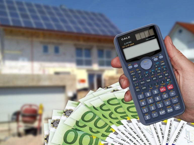 Hand hält Taschenrechner, dahinter ein Fächer mit 100 € Scheinen und im Hintergrund ein Haus, welches sich im Bau befindet