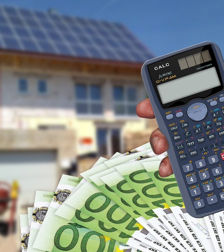 Hand hält Taschenrechner, dahinter ein Fächer mit 100 € Scheinen und im Hintergrund ein Haus, welches sich im Bau befindet