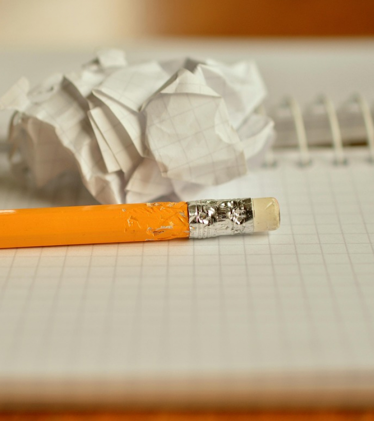 Bleistift mit Radiergummi liegt auf einem karierten Notizblock mit Spirale. Hinter dem Bleistift ein zusammengeknülltes Papier