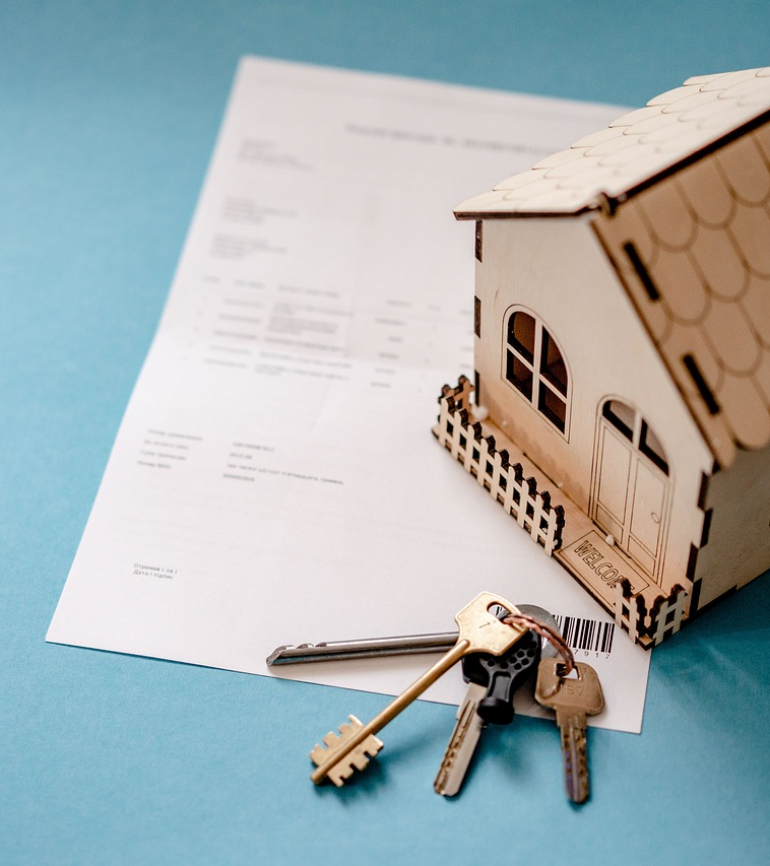 Blatt Papier mit Minihaus und Schlüssel liegen auf blauem Untergrund
